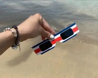 Hand holding colorful red, white, and blue striped solar eclipse glasses at a beach in Costa Rica, highlighting the design using the colors of the flag of Costa Rica.