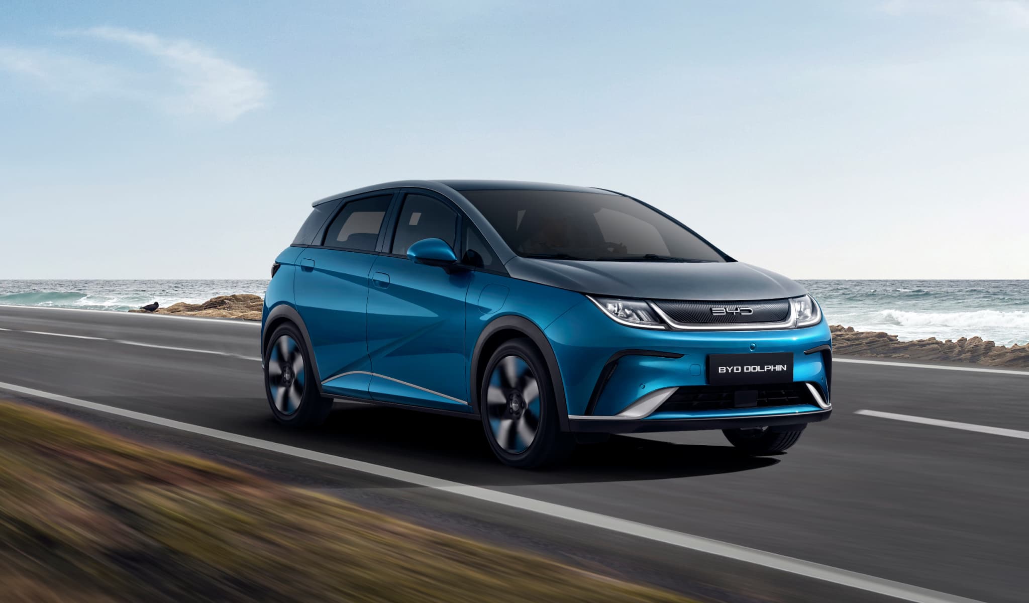 Blue BYD Dolphin electric car driving along a coastal road under clear sky